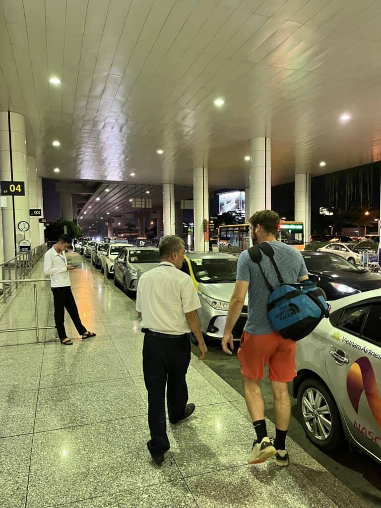Airport pickup area at night with a row of parked taxis under a well-lit canopy. A traveler with a blue North Face duffel bag walks alongside an airport staff member. Another person stands near the curb, checking their phone, while buses and additional vehicles are visible in the background.