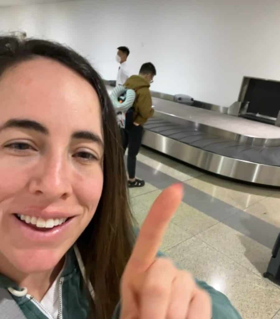 Woman smiling and pointing toward an empty baggage carousel in an airport. Behind her, two people stand near the carousel, one carrying a coiled neck pillow. The scene suggests a waiting moment in the luggage claim area.