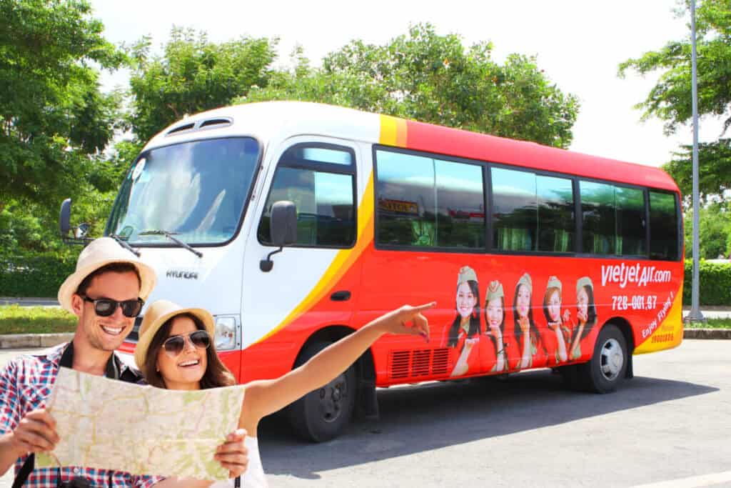 "A smiling couple holding a map stands in front of a red VietJet Air shuttle bus. The bus features branded graphics with airline hostesses and the VietJet Air logo, parked against a backdrop of lush green trees. The couple appears to be planning their next move, enjoying a sunny day."